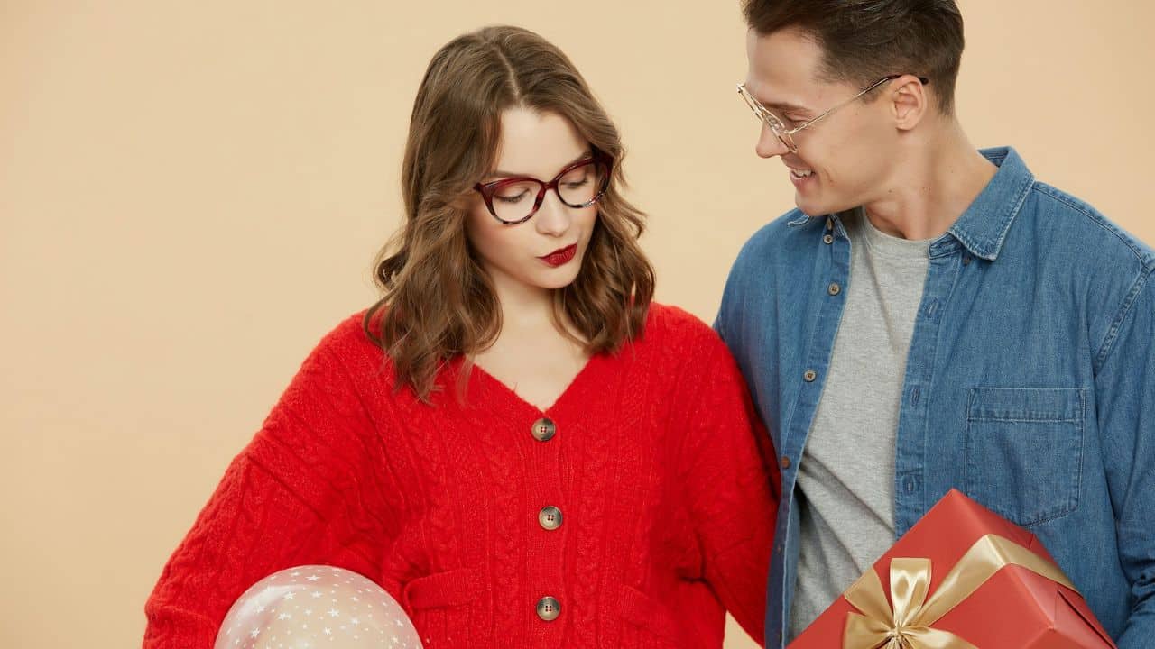 A woman in a red sweater looks down at a gift, while a man smiles at her, holding a large wrapped gift box.