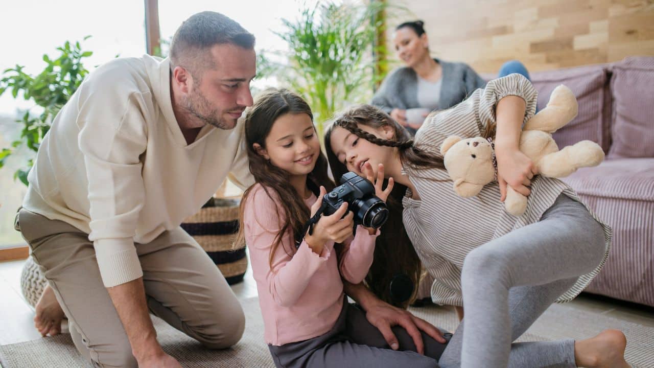 A father and his two daughters are on the floor looking at a camera, with the mother watching them from the couch.