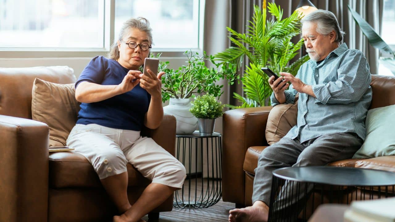 An older couple sits on separate armchairs, both looking at their phones.