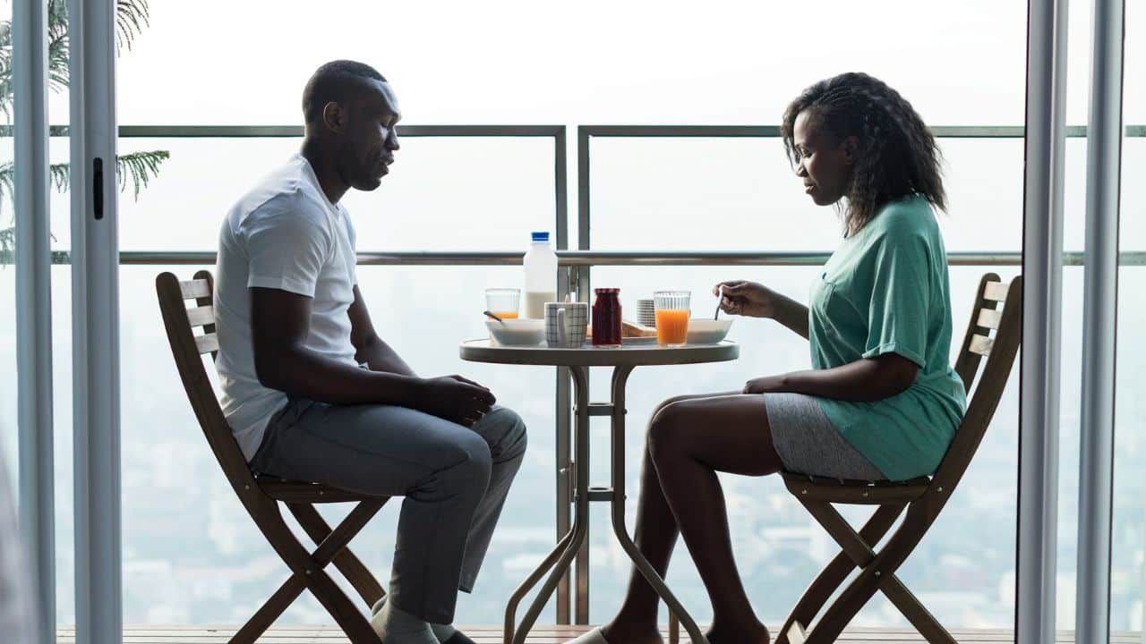A man and a woman sit facing each other at a small table on a balcony, not speaking.