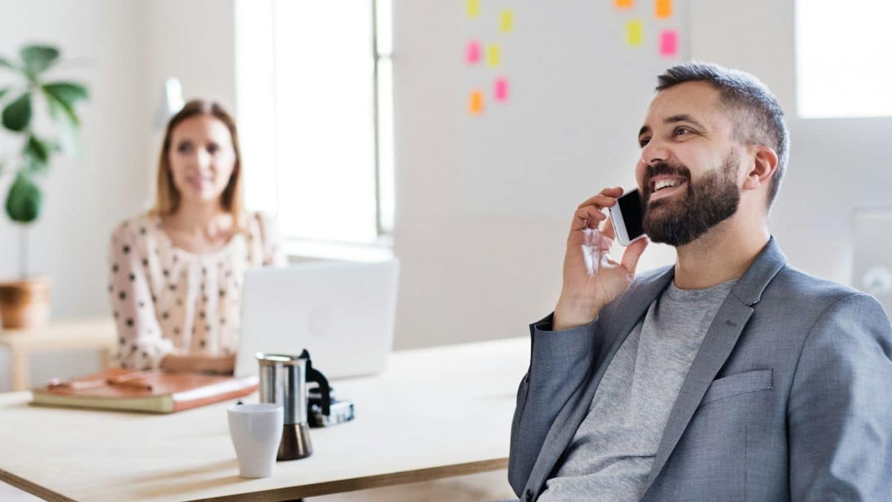 A man in an office smiling while talking on a phone, with a woman in the background.