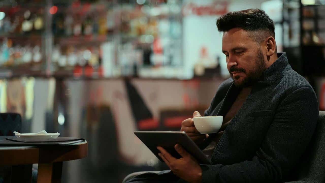 A handsome man sits alone in a restaurant or bar, holding a cup of coffee and looking at a tablet.