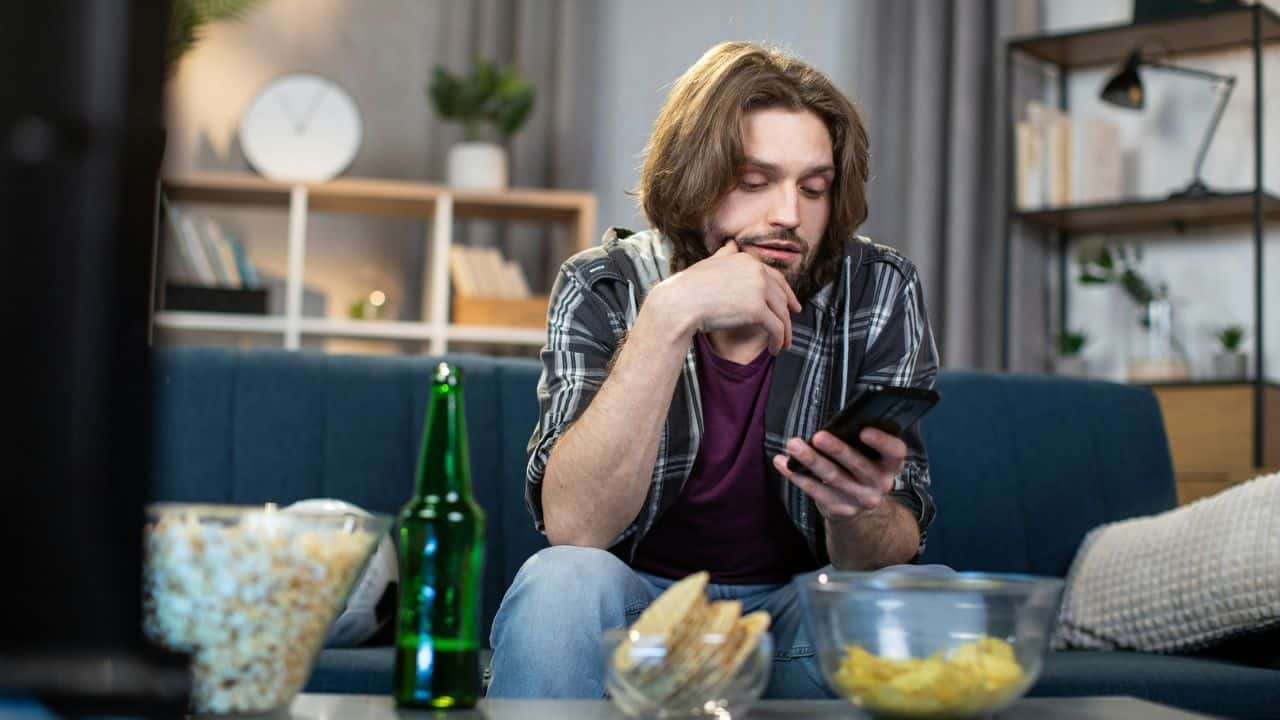 A man sits on a couch, looking at his phone with a beer, popcorn, and chips on the coffee table.