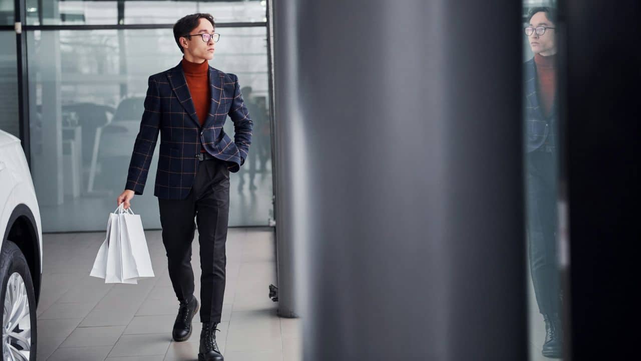 A fashionable man in a blazer and a turtleneck, carrying shopping bags, walks past a car showroom window.