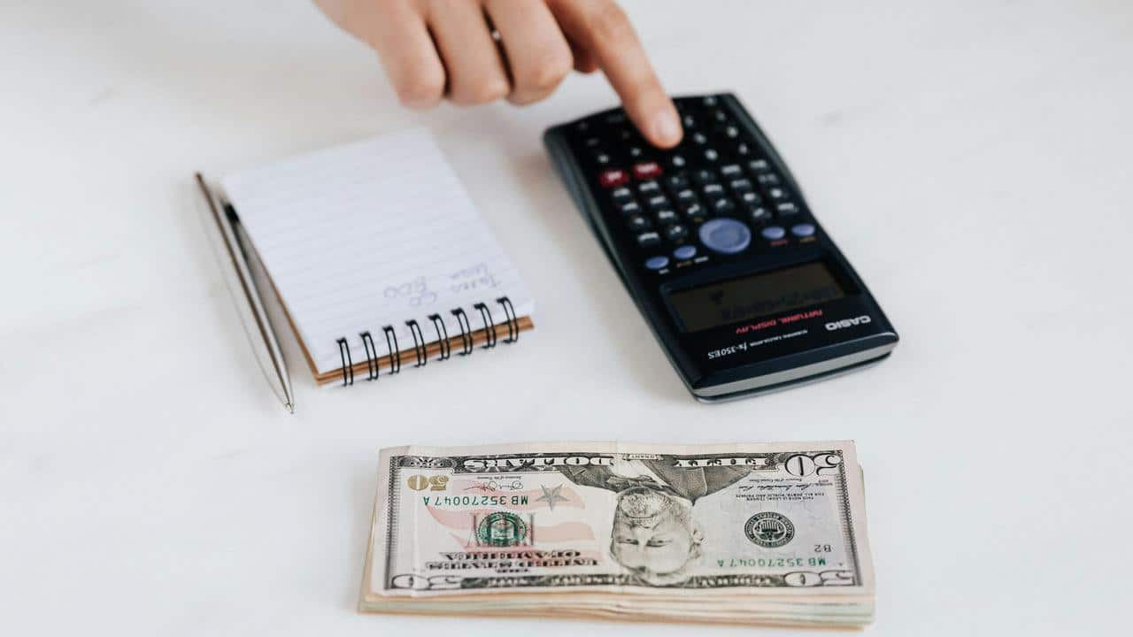 A person’s hand using a calculator beside a notepad and a stack of fifty-dollar bills.