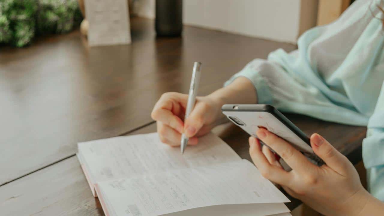 A person writing in a notebook while holding a smartphone.