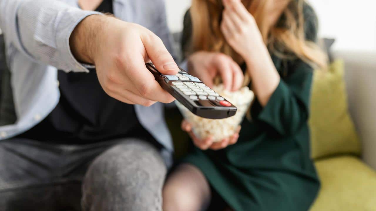 A person holding a TV remote while another eats popcorn.