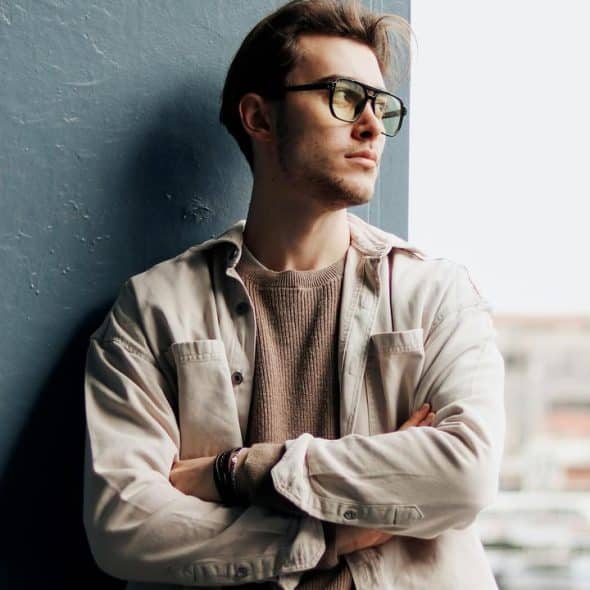 A man in glasses leans against a wall with arms crossed.