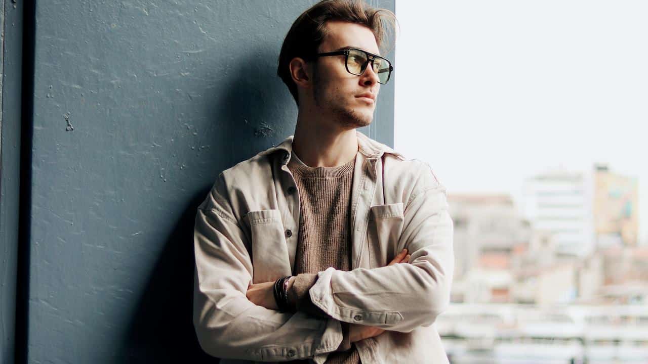 A man in glasses leans against a wall with arms crossed.