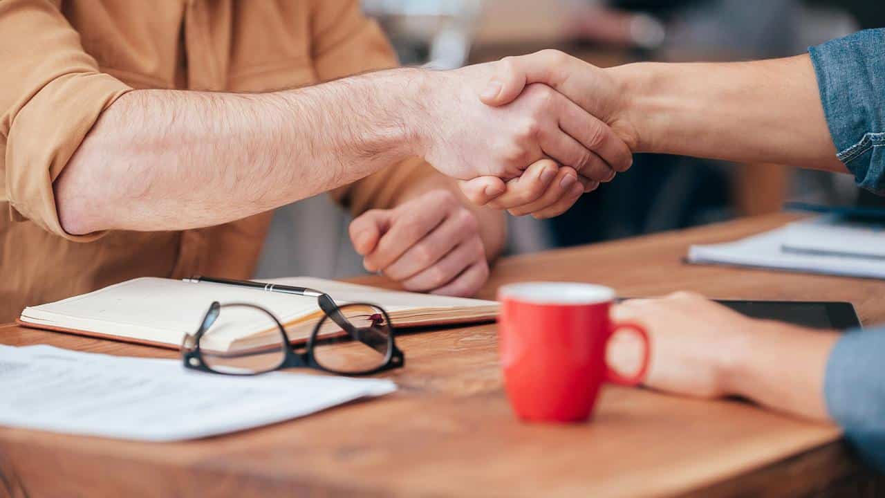 A close-up of two people shaking hands over a desk with papers and a notebook.