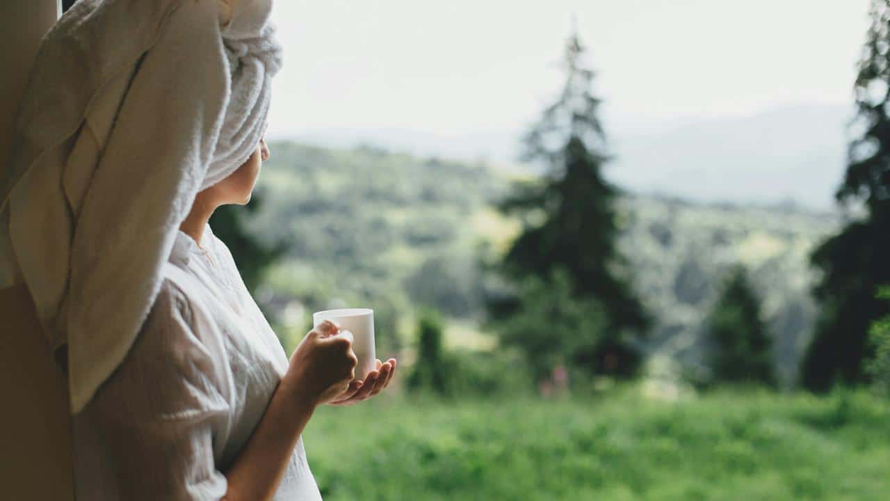 A person with a towel on their head holding a mug and looking outside.