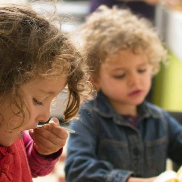 A girl eating while another child peels a banana.