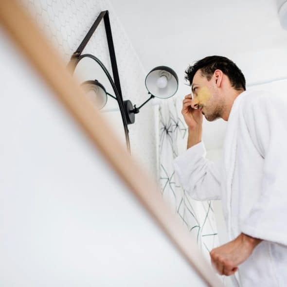 A man in a bathrobe applying a face mask in front of a mirror.