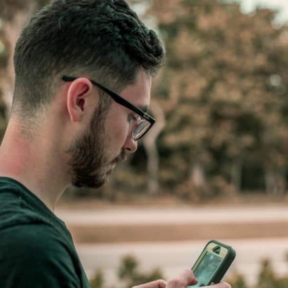 A bearded man with glasses looking at his smartphone outdoors.