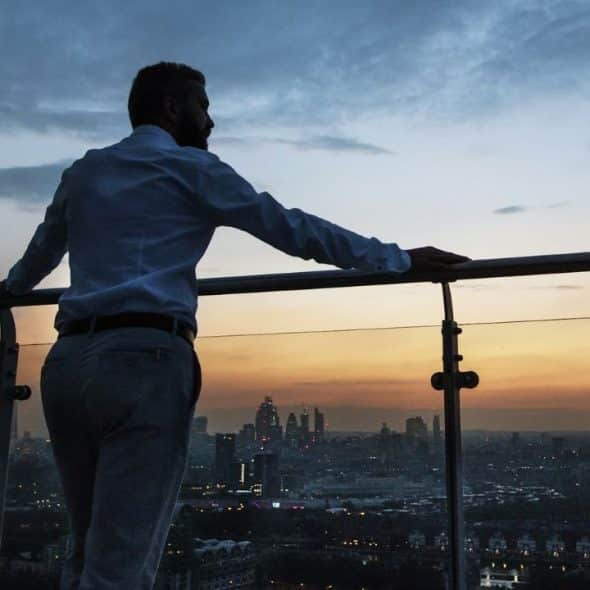 A man in a button-down shirt stands on a balcony overlooking a city at dusk.