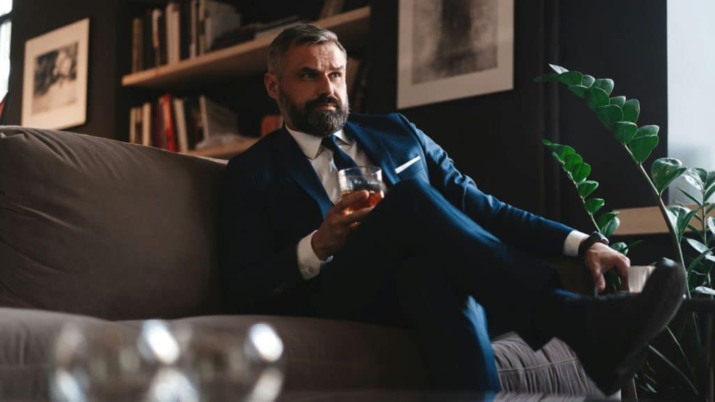A handsome, middle-aged man with a beard sits on a couch, holding a drink and looking thoughtfully away from the camera.