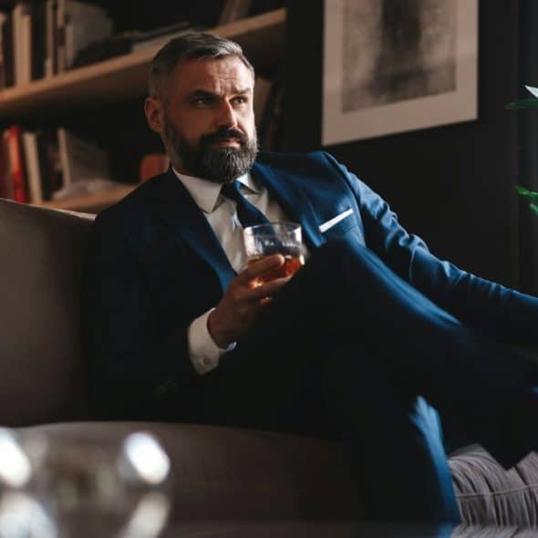 A handsome, middle-aged man with a beard sits on a couch, holding a drink and looking thoughtfully away from the camera.