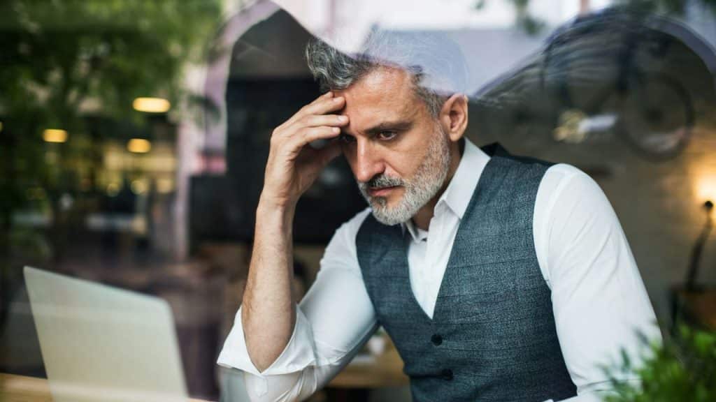 A distressed, gray-haired man in a waistcoat sits at a table with his head in his hand, looking down at a laptop.