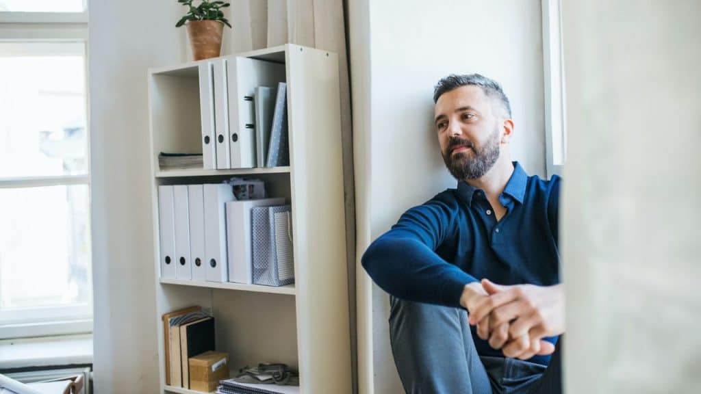 A thoughtful man sits on the floor next to a bookshelf, with his hands clasped and a distant look.