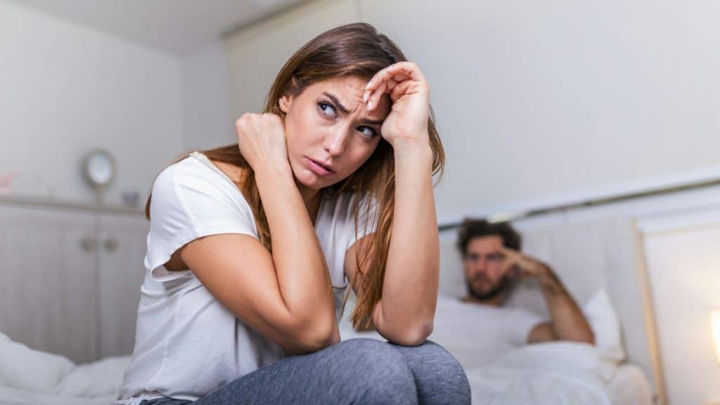 A sad and worried woman sits in the foreground, while a man in the background is out of focus.