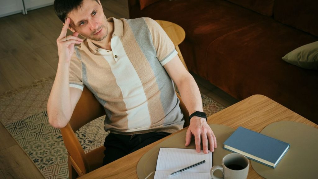 A man sits at a table, looking away in deep thought with his hand to his head.