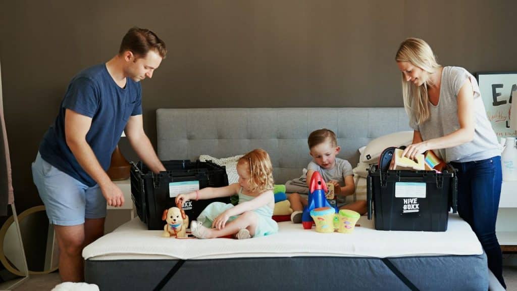 A family is in a bedroom, with two parents and two children putting toys in black storage bins on a mattress.