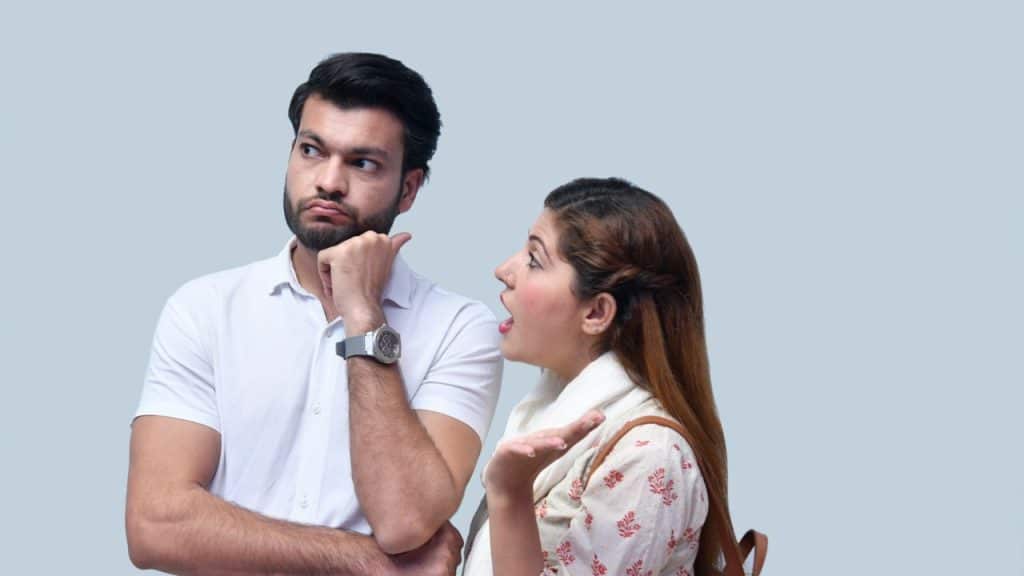 A man with his arms crossed looks away from a woman, who is speaking and gesturing with a frustrated expression.