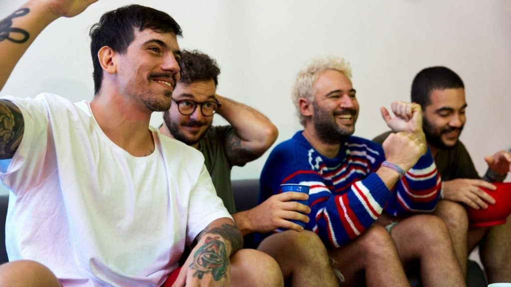 A diverse group of men sit on a couch, smiling and cheering while watching something.