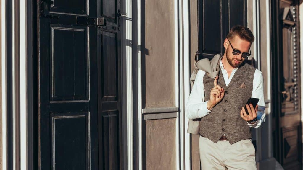 A man in sunglasses with a jacket over his shoulder checking his phone.