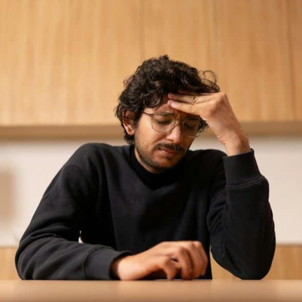 A man with curly hair and glasses sits at a table, holding his head in one hand.