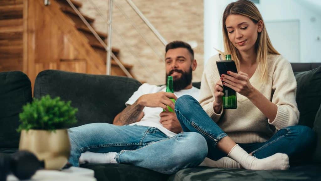 A couple relaxing on a couch with beer bottles as the woman checks her phone.
