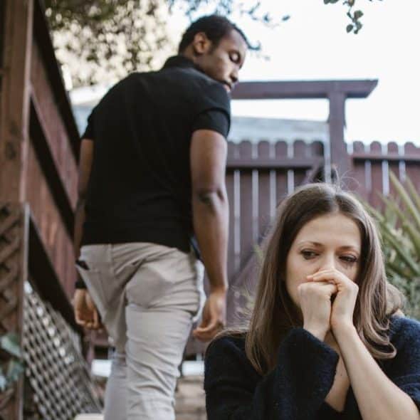 A woman is looking down with her hands near her face, and a man is walking away in the background.