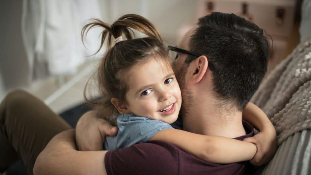 A smiling little girl with her hair in a ponytail is hugging a man.