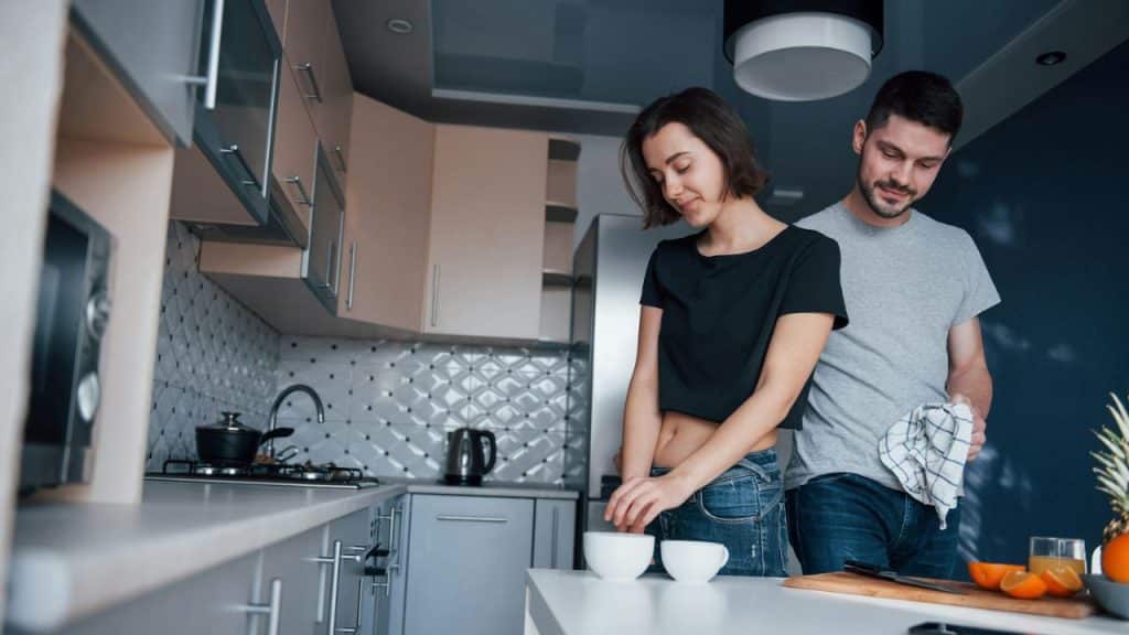 A woman and a man are standing close to each other in a kitchen. The woman is reaching for a mug, and the man is drying a plate with a towel.