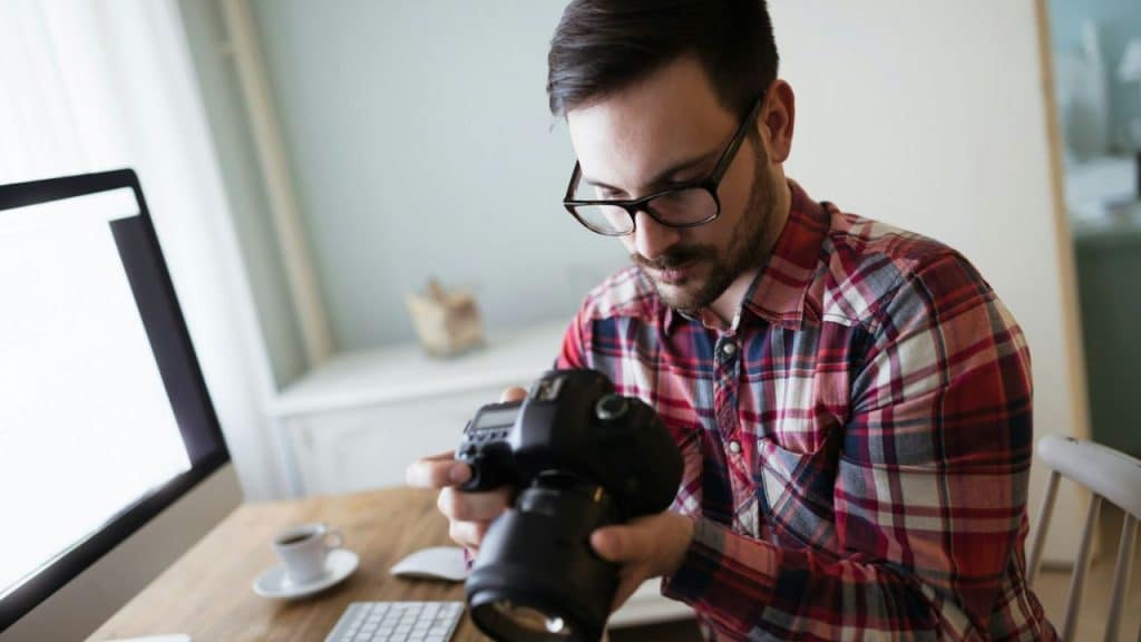 A man with glasses and a plaid shirt is looking at a camera while sitting at a desk.