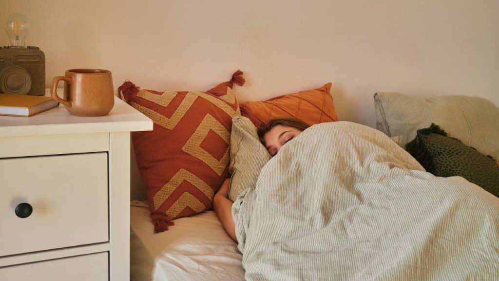 A person is lying in bed, covered by a blanket, with their eyes closed. A nightstand with a mug and book is next to the bed.