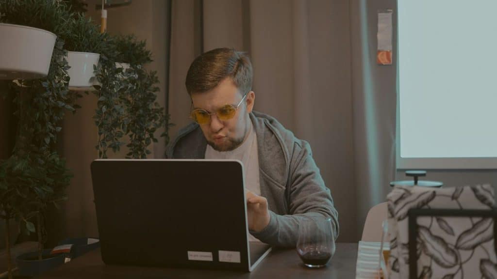 A man with a beard, yellow sunglasses, and a gray hoodie is looking at a laptop. There is a glass of red liquid on the table next to him.