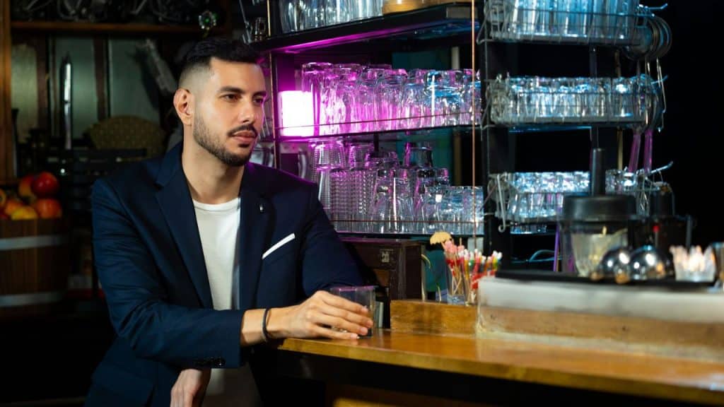 A man in a blue blazer and white shirt is sitting at a bar with a glass in his hand.