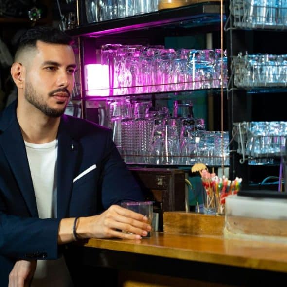 A man in a blue blazer and white shirt is sitting at a bar with a glass in his hand.