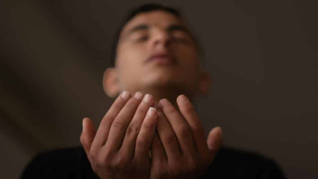 A blurry figure with their eyes closed is holding their hands out, palms up.