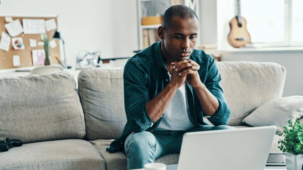 A man is sitting on a couch, looking at a laptop with his hands clasped together. A guitar is visible in the background.