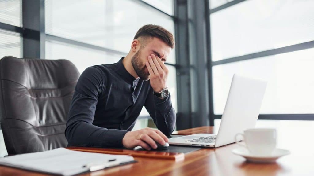 A tired man in a black shirt is rubbing his eyes at his desk.