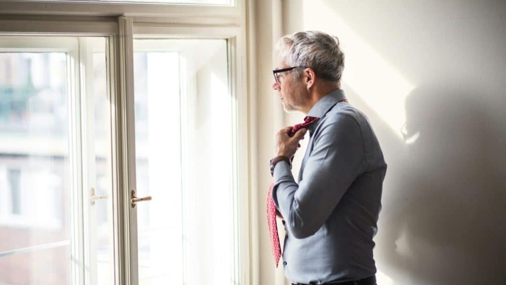 A man with gray hair and glasses is loosening his red tie while looking out a window.