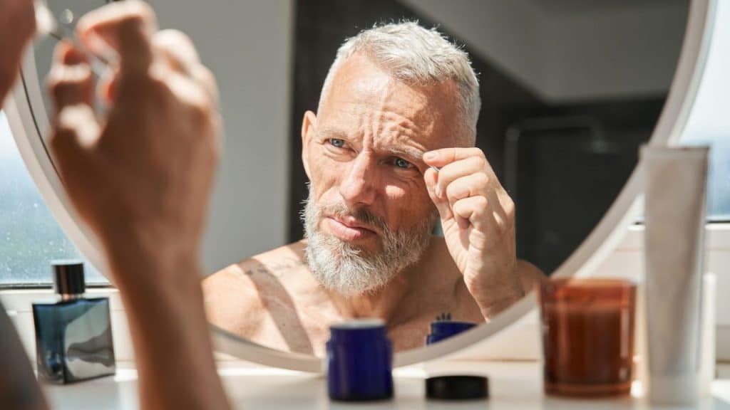 A man with a white beard and gray hair is reflected in a mirror while he plucks his eyebrow.