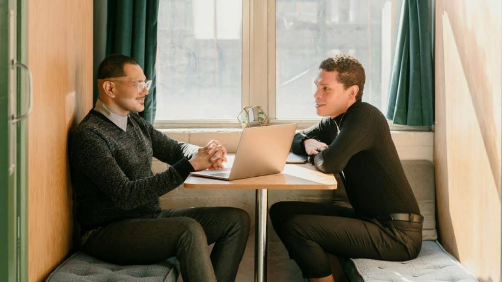 Two men are sitting across from each other at a small table with a laptop.