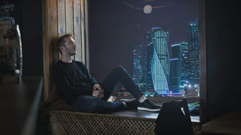 A man sits alone on a window sill looking at a city skyline at night.