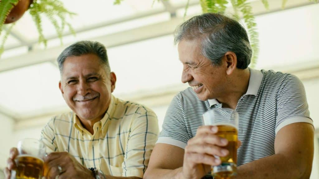 Two middle-aged men with gray hair sit and drink beers, looking at each other and smiling.