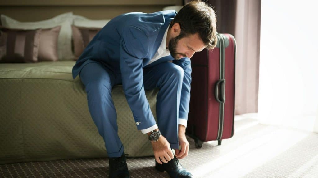 A man in a blue suit is sitting on a bed tying his shoes next to a suitcase.