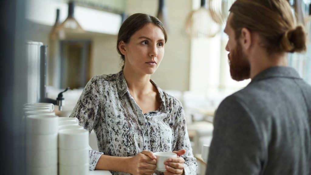 A woman holding a cup while talking to a man indoors.