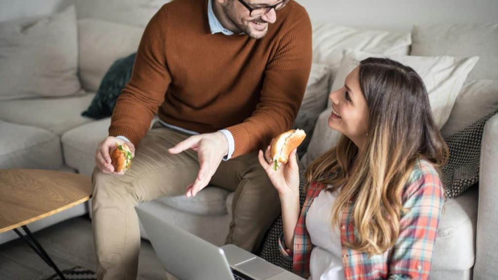 A couple eating sandwiches and talking while sitting near a laptop.